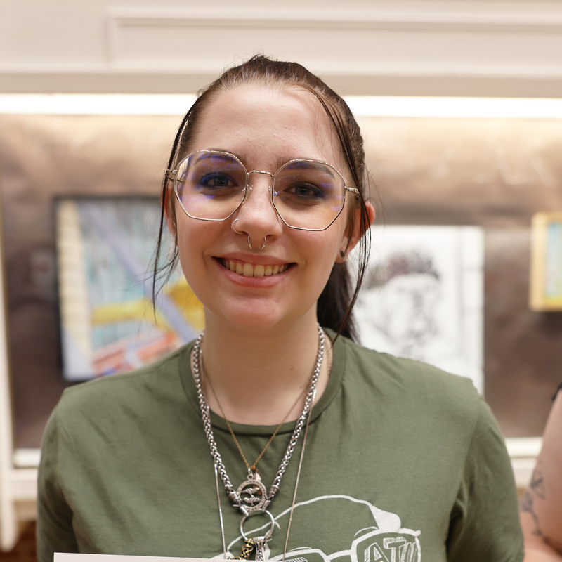 Young woman with black hair, a greenshirt, glasses, and a nose piercing smiles into the camera.